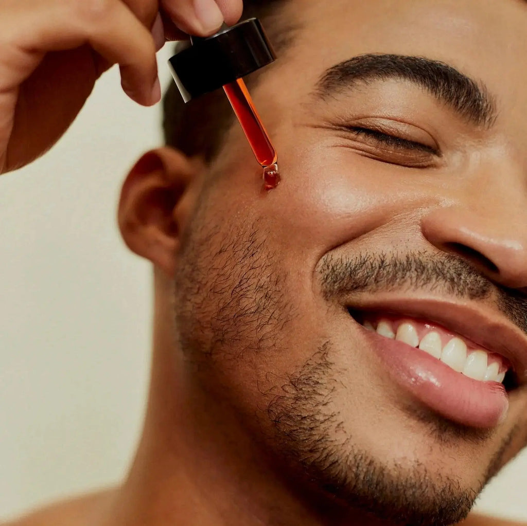 Close-up of a smiling man applying Love Me, Heal Me® Facial Serum to his cheek with a dropper. Healthy, glowing skin and pure self-care joy.