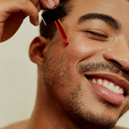 Close-up of a smiling man applying Love Me, Heal Me® Facial Serum to his cheek with a dropper. Healthy, glowing skin and pure self-care joy.
