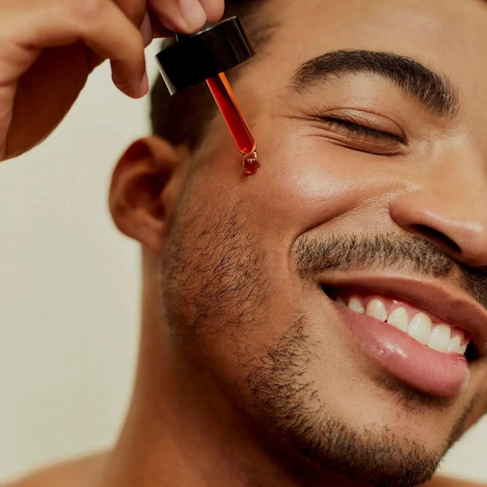 Close-up of a smiling man applying Love Me, Heal Me® Facial Serum to his cheek with a dropper. Healthy, glowing skin and pure self-care joy.