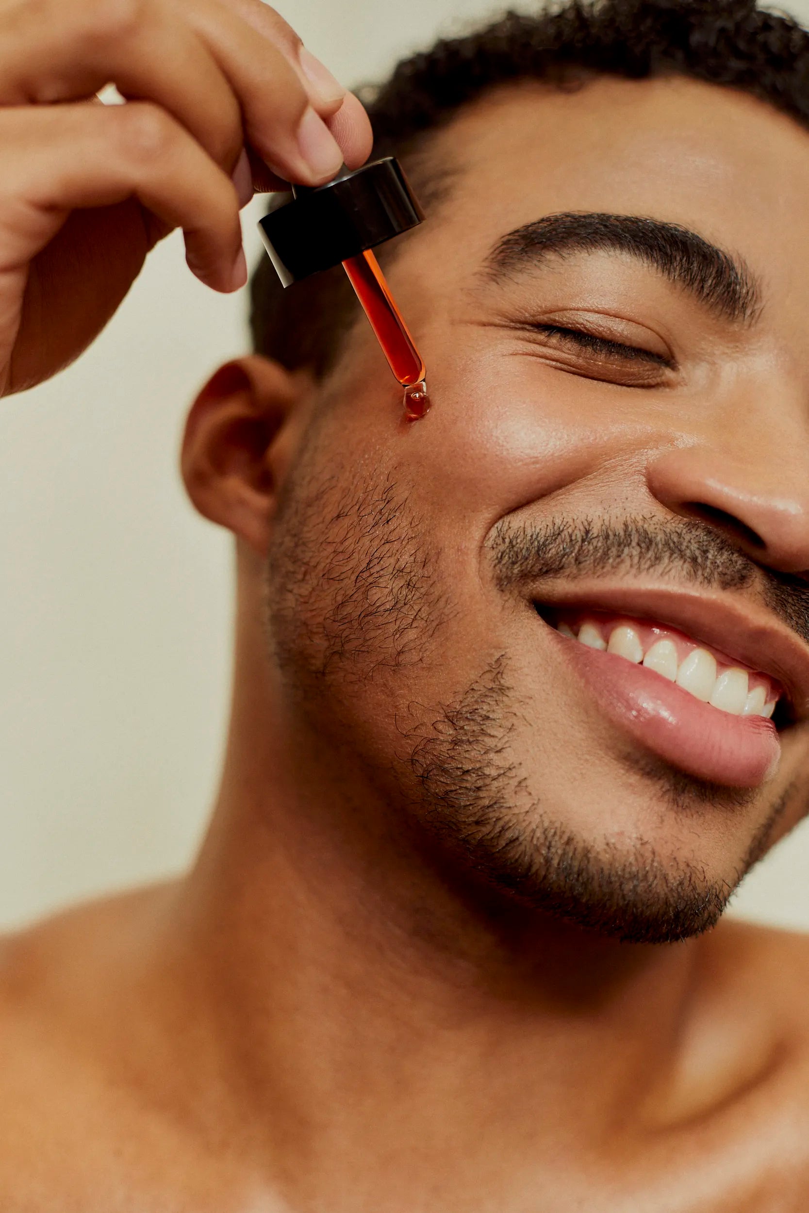 Close-up of a smiling man applying Love Me, Heal Me® Facial Serum to his cheek with a dropper. Healthy, glowing skin and pure self-care joy.